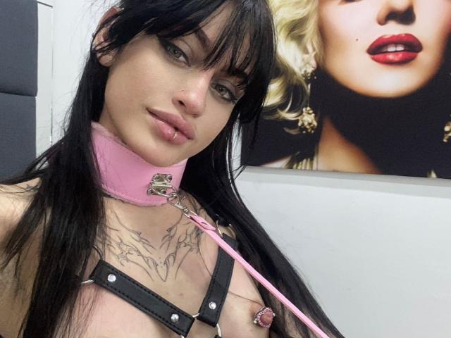 SofiaBlossom with black hair, pink collar, black leather harness, tattoos on chest, pink leash, red lipstick, blonde photo in background. Boobs, collar, tattoos, leash visible.