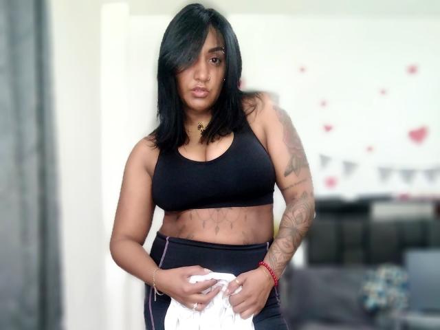 Nahirr has medium brown skin, black shoulder-length hair, wearing a black sports bra showing her tits and black pants, holding white clothes, tattooed arm, red bracelet.