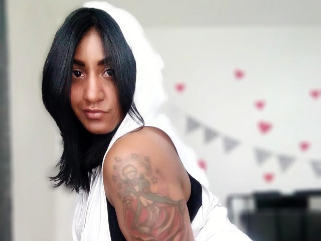 Nahirr with shoulder tattoo of a woman holding a sword, black hair, white sleeveless top, nose ring, and a subtle smile, looking over shoulder.
