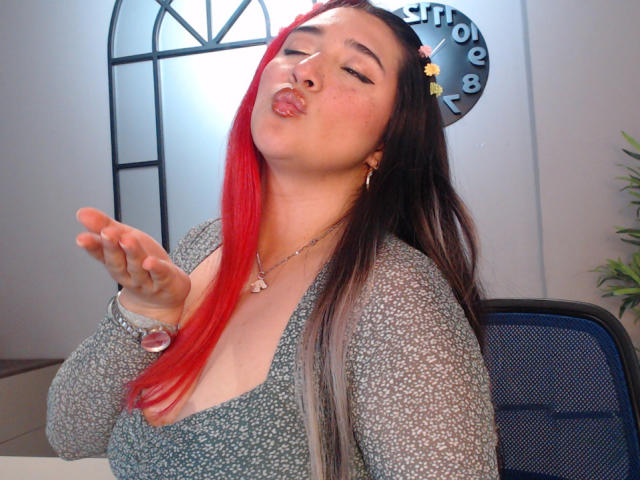 IsabellaClown with red and black hair, puffy lips, wearing a grey patterned top, silver necklace, and earrings, making a "shush" gesture with a finger to her lips.