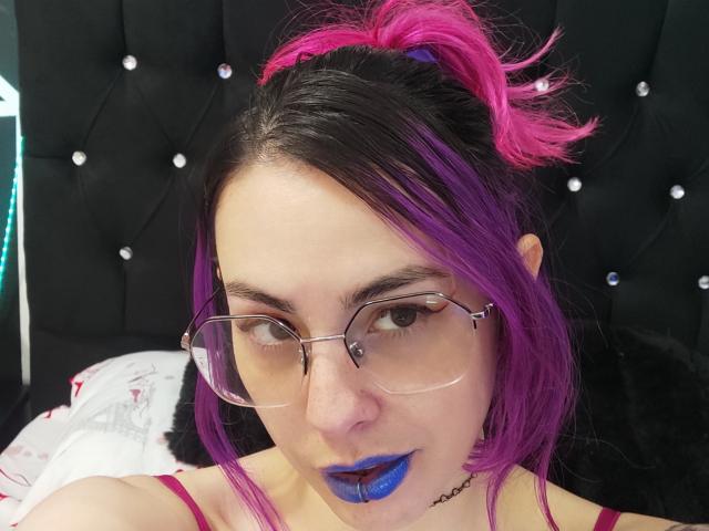 NatashaLau with purple and pink hair in a ponytail, wearing glasses, blue lipstick, and a black choker, looking at the camera, against a black tufted headboard.