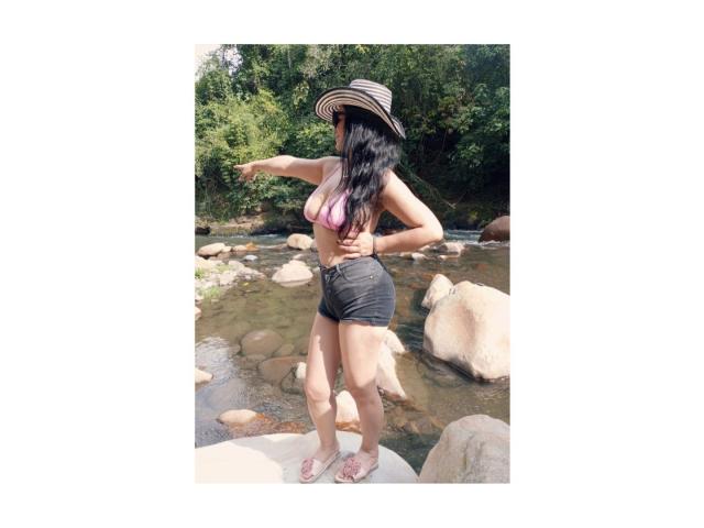 CameronViviane stands in a rocky creek, wearing a pink bikini top, black shorts, and a striped hat, with long black hair, pointing forward, barefoot, in sunlight.