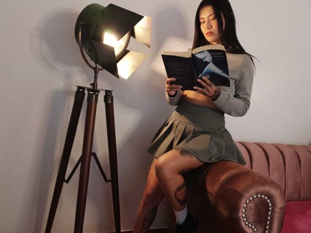 AsshleyCollins with long black hair, wearing a gray crop top and green skirt, reading a book on a brown leather couch, next to a standing lamp with a bright bulb.