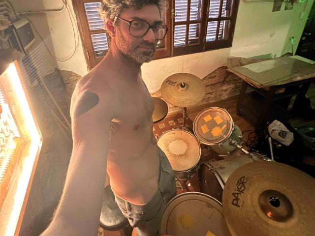 Shirtless Kaik with glasses, beard, and tattoo on left shoulder, standing in front of drum set, wearing gray pants, in dimly lit room, holding phone for selfie.