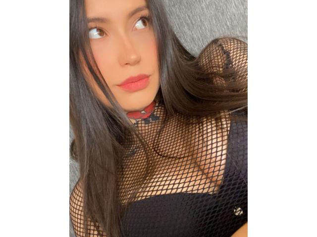 Asshley Collins, con cabello largo y oscuro, vestida con una blusa negra de rejilla, mostrando sus pechos, y un collar rojo, mirando hacia un lado con lápiz labial rojo en sus labios carnosos..