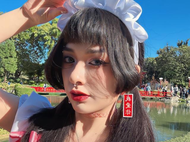 SabriPrettty, con cabello oscuro en coletas, diadema blanca de sirvienta, lápiz labial rojo, delineador de ojos negro, vestida con un conjunto rojo y blanco, de pie junto a un estanque, cielo azul claro..