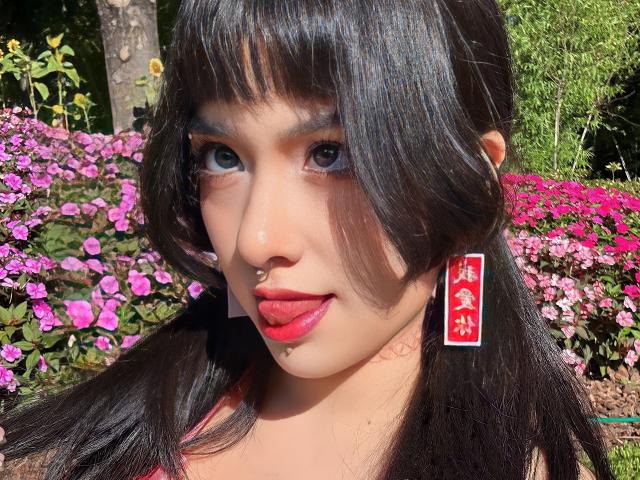 SabriPrettty a les cheveux noirs et brillants, porte du rouge à lèvres et un anneau de nez. Elle porte des boucles d'oreilles rouges avec des caractères chinois blancs et elle tire la langue..