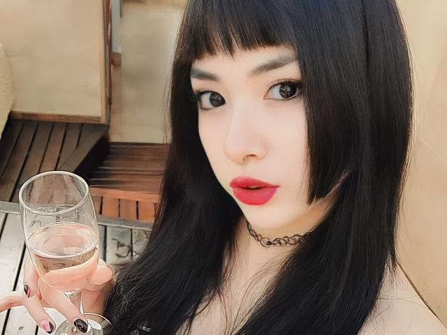 SabriPrettty, con cabello negro y liso, lápiz labial rojo y un collar negro, sosteniendo una copa de vino rosado, mirando a la cámara con grandes ojos oscuros, mostrando ligeramente sus senos..