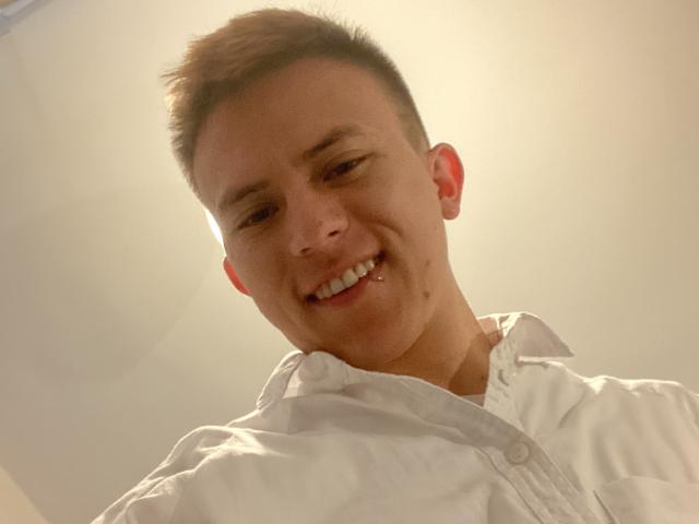 BastianC, sorridendo con un solo dente visibile, indossa una camicia bianca, ha i capelli castani corti e una piccola lentiggine sulla guancia destra, in un selfie scattato da un'angolazione bassa..