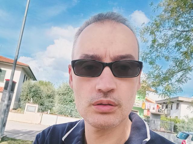 Tarabas, met kort, grijs haar, zwarte zonnebril en een donker poloshirt, heeft een serieuze, licht geïrriteerde uitdrukking en een lichte baard, op een daglichtfoto die hij zelf heeft gemaakt..