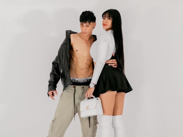 KathyAndZac, avec des cheveux noirs, un haut blanc, une jupe noire, des bottes blanches, tenant un sac à main, se tient à côté de Zac, qui a les cheveux bouclés, est torse nu, porte une veste noire et un pantalon gris, et montre son entrejambe..