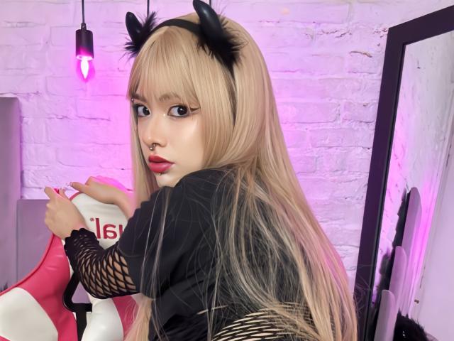 SabriPrettty met lang blond haar, zwarte kattenoren, zwarte mesh mouwen en rode lippenstift, kijkt over haar schouder, terwijl ze een roze en witte stoel vasthoudt, met een neusring..