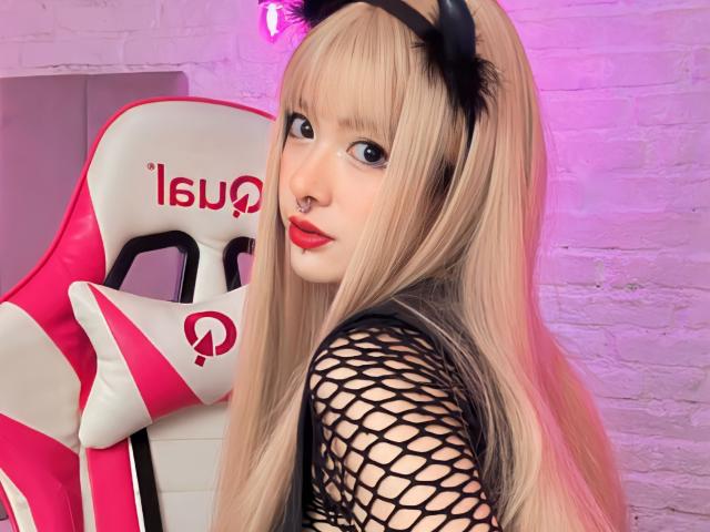 SabriPrettty met lang blond haar, een zwarte top van netstof, een hoofdband met kattenoren, rode lippenstift, een neusring, kijkt over haar schouder en zit in een roze en witte gamingstoel..