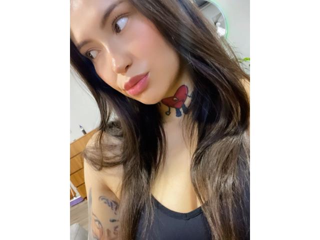 Asshley Collins a de longs cheveux foncés, des lèvres roses et un tatouage en forme de cœur rouge sur son cou. Elle porte un haut noir et regarde sur le côté avec un léger sourire..