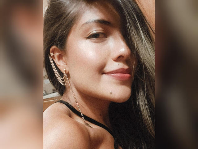 SunriseX, con lunghi capelli castani, indossa un top nero con spalline e ha una piccola lentiggine sulla guancia destra e un orecchino a catena, sorridendo dolcemente. La sua pelle è abbronzata e liscia..