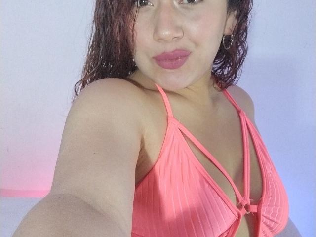 VeronicaHot69, com cabelo castanho úmido, biquíni listrado de rosa, pequenos brincos de argola dourados e batom rosa, mostrando seus seios em um selfie. Ela está com uma expressão levemente emburrada..