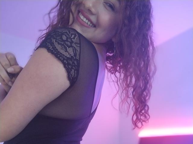 VeronicaHot69 met krullend rood haar, lachend, draagt een zwarte kanten top, toont haar rug en houdt haar arm. Haar borsten zijn zichtbaar door het dunne kant..