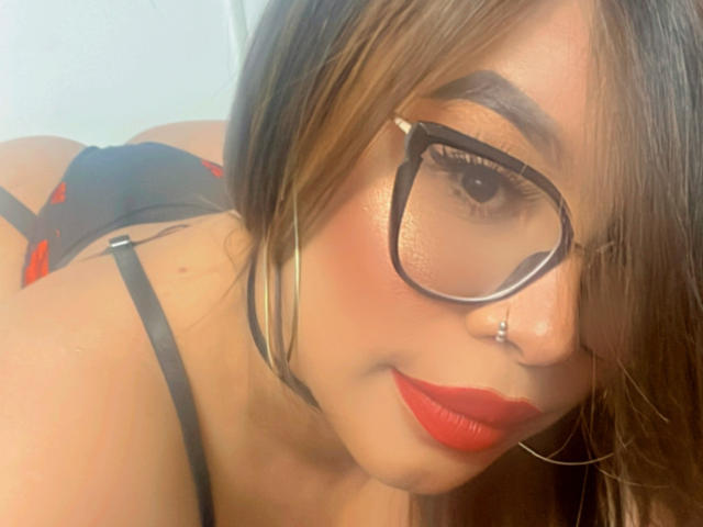 ValeriHarris con gli occhiali, il rossetto rosso e il piercing al naso, indossa lingerie nera con dettagli rossi, mostrando il sedere, i capelli castani e gli orecchini a cerchio in un selfie..