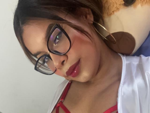 ValeriHarris, con gli occhiali, il rossetto rosso e gli orecchini a cerchio, indossa una camicia bianca e un reggiseno rosso, ha la pelle marrone chiaro e i capelli castano scuro, e sorride leggermente..