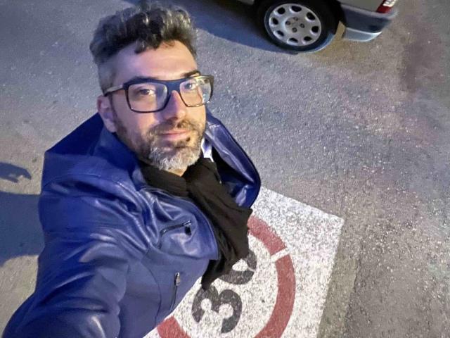 Kaik, mit lockigem, grauem Haar, Bart und Brille, trägt eine blaue Lederjacke und einen schwarzen Schal und macht ein Selfie auf einer Straße. Sein Hintern ist teilweise sichtbar..