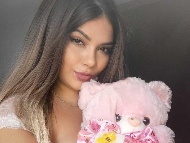 Atenaa, com cabelos castanhos longos, vestindo uma blusa branca de renda, segurando um urso de pelúcia rosa com olhos florais, olhando para a câmera com um leve sorriso..