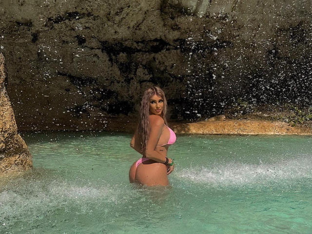 TanyaMonroe con un bikini rosa, con agua salpicando alrededor de su trasero, de pie en agua verde cristalina con una cueva rocosa detrás de ella. Cabello largo de color rosa parduzco, piel bronceada, sonriendo..