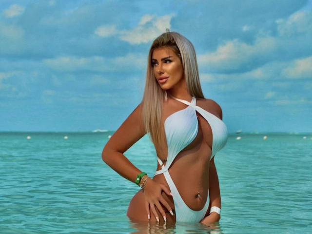 TanyaMonroe con un busto grande, vestida con un traje de baño blanco de una sola pieza con cortes, de pie en agua azul cristalina, mirando a la cámara, con pulseras verdes y mostrando un piercing en el ombligo..