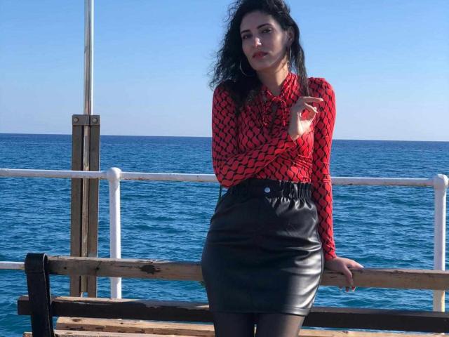 AddictedToUTS con i capelli neri ricci, una camicia rossa a motivi e una gonna di pelle nera, in piedi vicino a una ringhiera di legno, con una sigaretta in mano, l'oceano blu sullo sfondo, cielo sereno, indossa collant neri..