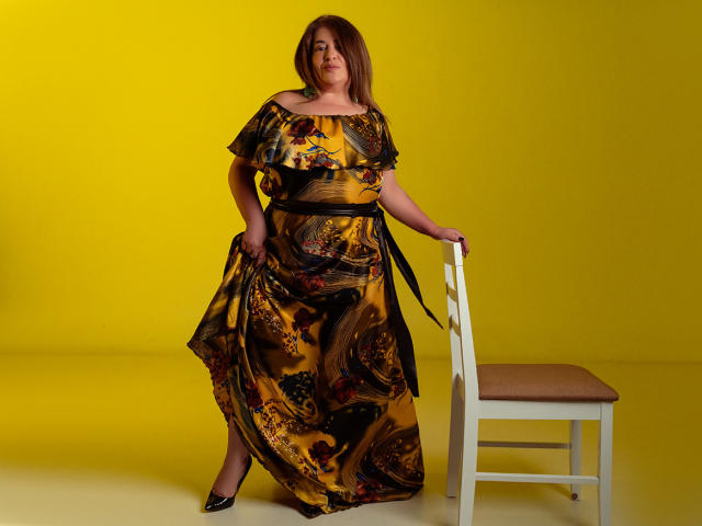 Marissa Serano steht mit einer Hand auf einem weißen Stuhl, trägt ein gelb-schwarzes Blumenkleid, schwarze Absätze und einen Gürtel, vor einem hellgelben Hintergrund..