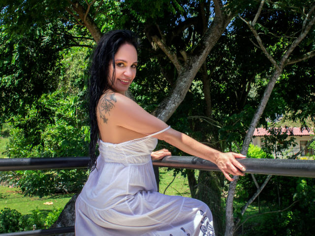 Laila Castillo, avec de longs cheveux noirs, souriante, portant une robe blanche sans bretelles, montrant son épaule tatouée, assise sur une rambarde noire, avec des arbres verts en arrière-plan, une journée ensoleillée..