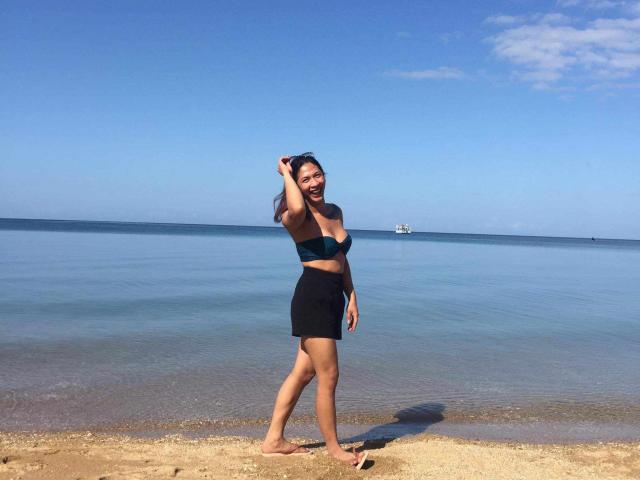 SexyFantasyYuri standing on a sandy beach, wearing a black strapless top and black skirt, with her right hand in her hair, smiling, barefoot, and looking happy.