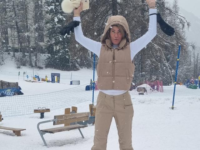 BlackAngeel stands in a snowy park, arms raised, wearing a beige puffy vest, beige pants, white long-sleeve shirt, and beige hat, holding white gloves, smiling.