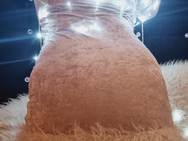 Il sedere di CatalinaJH in un abito rosa chiaro con una texture particolare, una sovrapposizione di plastica trasparente, luci bianche che brillano sullo sfondo e un tappeto bianco soffice in basso..