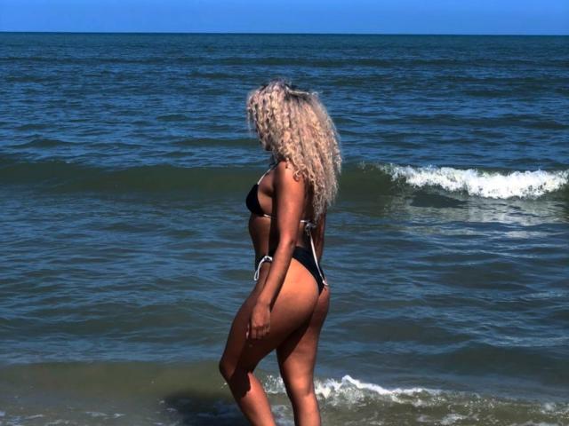 LilithWalker with curly blonde hair in a black bikini, standing in shallow ocean water, showing her ass and back, looking to the side.