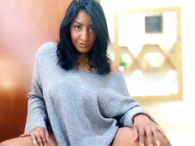Nahirr with medium brown skin and black wavy hair, wearing a loose gray sweater, sitting with legs apart, showing her ass, and a subtle smile, in a softly lit room.