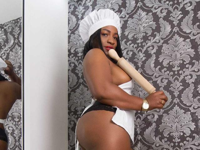 RousJunes, di carnagione scura con lunghi capelli neri, indossa una cuffia da cameriera bianca, tiene un mattarello di legno contro il seno, con indosso mutandine di pizzo nero, mostrando il suo grande sedere..