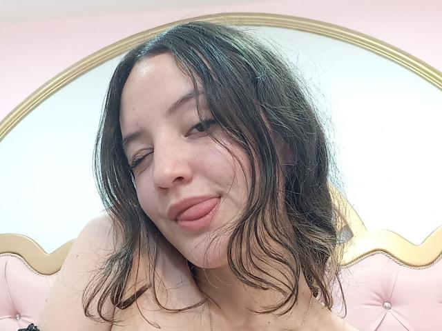 ChloeDulce, con el pelo castaño despeinado, guiñando un ojo y sacando la lengua de color rosa. Está sin ropa, mostrando sus hombros. Tiene la piel clara y una expresión linda y juguetona..