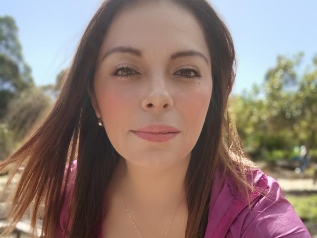 El rostro de Seher, con piel clara, ojos marrones y cabello largo castaño, vestida con una camisa rosa, con una pequeña marca de nacimiento sobre su labio superior derecho, y un collar de oro..