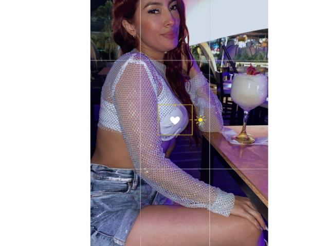 KendraMiler with red hair, wearing a see-through mesh top and denim shorts, sitting at a bar, her tits visible, holding a cocktail, looking at the camera.