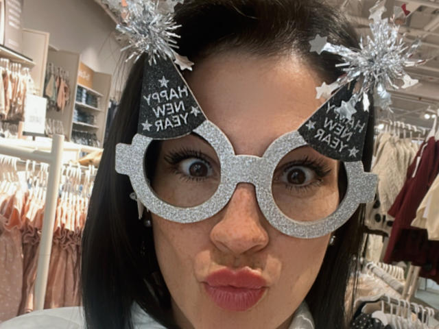 Liliysweet wearing glittery silver party hat with "Happy New Baby" text and sparkly pom-poms, puffy pink lips, black hair, and wide brown eyes, in a store.