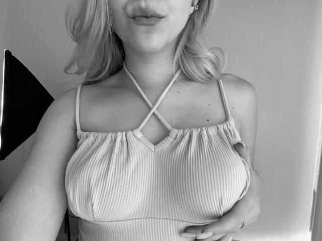 Black and white photo of SugarrX with blonde hair, wearing a ribbed, criss-cross halter top, hand on her tits, soft smile, visible earrings.