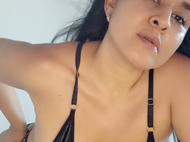 Clariveth mit dunklem Haar, die einen schwarzen Leder-Bikinitop und -Gürtel trägt, wobei ihre großen Brüste sichtbar sind und sie ein selbstbewusstes Gesicht zeigt, leicht nach vorne gebeugt. Sie hat ein leichtes Lächeln..