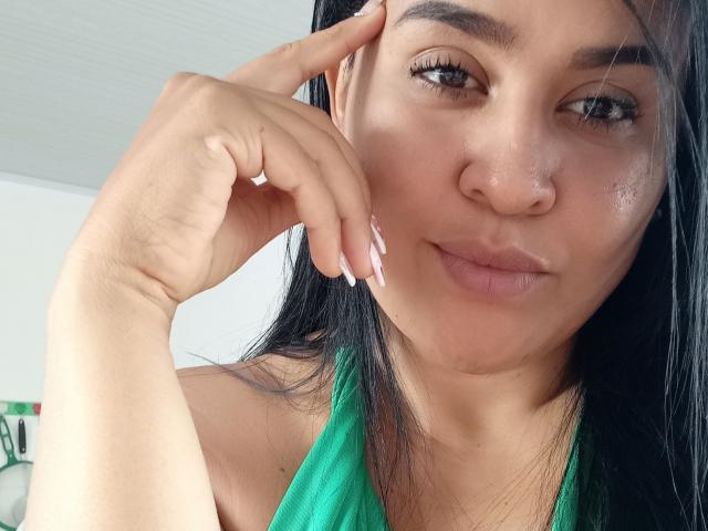 Clariveth, met donker haar, bruine ogen en lichtbruine huid, raakt haar gezicht aan met haar rechterhand, draagt een groene haltertop, waardoor haar kleine borsten en roze nagels zichtbaar zijn..