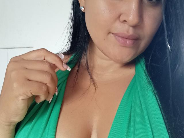 Clariveth, con piel morena, cabello negro y un vestido verde, lo sostiene abierto para mostrar sus pechos. Tiene una sonrisa encantadora, pequeños aretes y está tocándose el cabello..