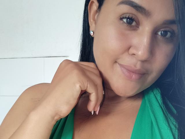 O rosto de Clariveth, blusa verde, cabelo preto, olhos castanhos, sorriso discreto, unhas cor de rosa, brinco de diamante, mão perto do pescoço, pele clara, maquiagem suave, blush discreto..