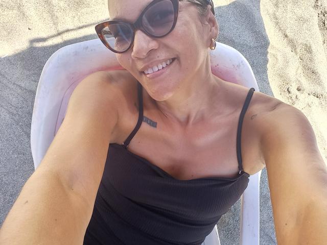 MatureJennyX with tan skin, black tank top, small tits, black sunglasses, earrings, smiling, sand, and sun, sitting on white chair, selfie, upper body, tattoos on chest.