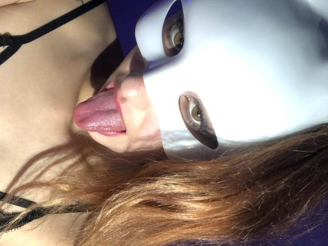 LilouSmoknu com uma máscara branca, língua para fora, olhos visíveis, cabelo castanho, alça preta, pele clara, língua rosa e uma leve sombra no rosto..