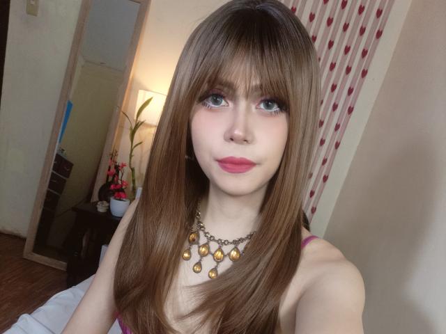 TSAnabelleSwift com cabelos castanhos longos, olhos azuis, batom rosa, usando um colar de ouro e uma blusa rosa, mostrando seus seios, em um quarto com um espelho e uma luminária..