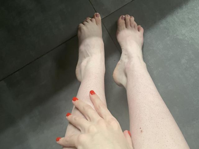 Les jambes pâles de RozalinaPleasure, parsemées de taches de rousseur, reposent sur des carreaux gris, avec du vernis à ongles rouge. Une main avec des ongles rouges touche son tibia, montrant ses orteils et ses doigts..