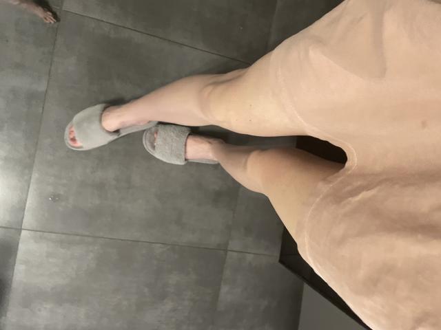 Las piernas desnudas de Rozalina, con pantuflas grises en los pies, de pie sobre un suelo de baldosas grises, vistiendo un vestido rosa transparente, con las puntas de los dedos pintadas de rojo..